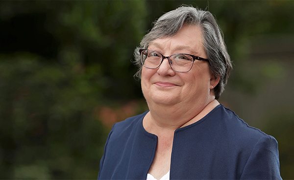 A smiling middle-aged woman, Cynthia Larive, in a blazer and glasses, standing in front of trees and flowers.